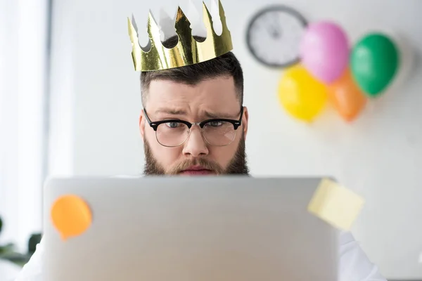 Portrait of businessman with paper crown on head looking at laptop screen in office — Stock Photo