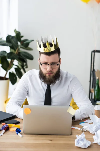 Porträt eines Geschäftsmannes mit Papierkrone auf dem Kopf, der am Arbeitsplatz im Büro auf dem Laptop tippt — Stockfoto