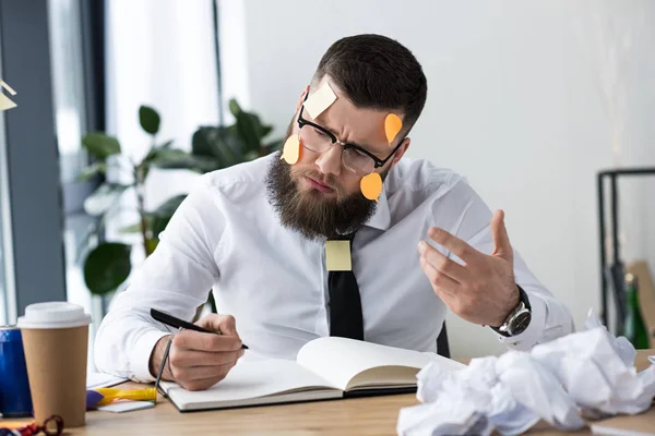 Portrait of businessman with sticky notes on face making notes in notebook at workplace — Stock Photo