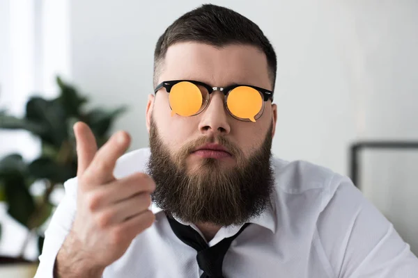 Retrato de hombre de negocios barbudo con notas adhesivas en las gafas apuntando a la cámara en la oficina - foto de stock