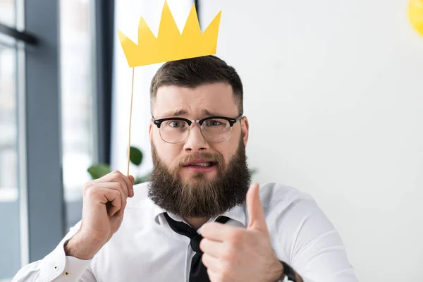 Portrait d'un homme d'affaires barbu avec couronne en papier montrant le pouce au bureau — Photo de stock