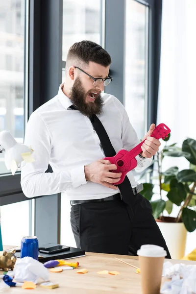 Эмоциональный бизнесмен, притворяющийся, что играет на игрушечной гитаре в офисе — стоковое фото