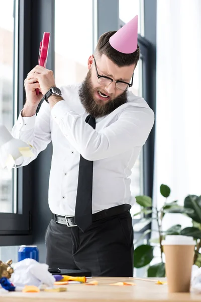 Businessman with party cone on head crashing toy guitar in office — Stock Photo