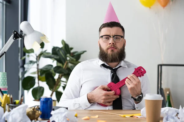 Portrait of businessman with party cone on head and toy guitar at workplace in office — Stock Photo
