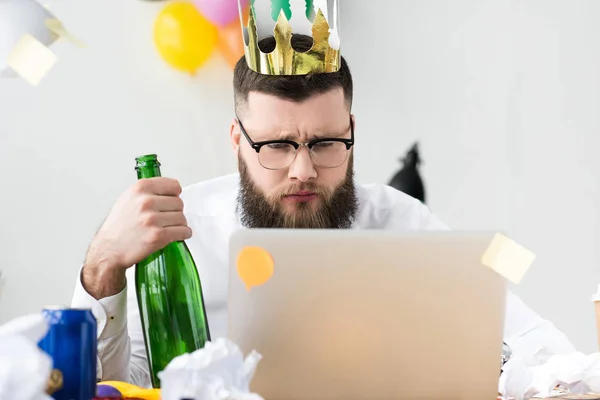 Empresário com coroa de papel na cabeça e garrafa de champanhe na mão olhando para a tela do laptop no local de trabalho — Fotografia de Stock