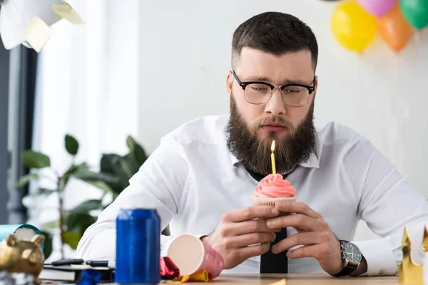 Portrait of sad businessman looking at birthday cupcake with candle in hands in office — Stock Photo