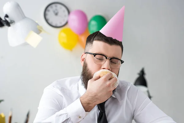 Ritratto di uomo d'affari in cono partito mangiare cupcake compleanno in ufficio — Foto stock