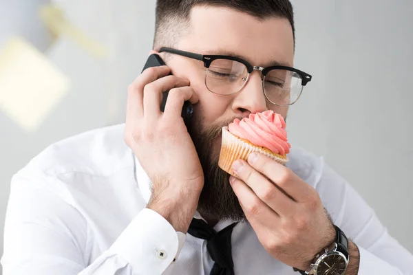 Retrato del hombre de negocios comiendo magdalena mientras habla en el teléfono inteligente - foto de stock