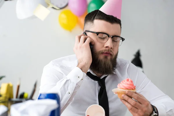 Porträt eines Geschäftsmannes in Partytüte mit Geburtstagskuchen in der Hand, der auf dem Smartphone spricht — Stockfoto