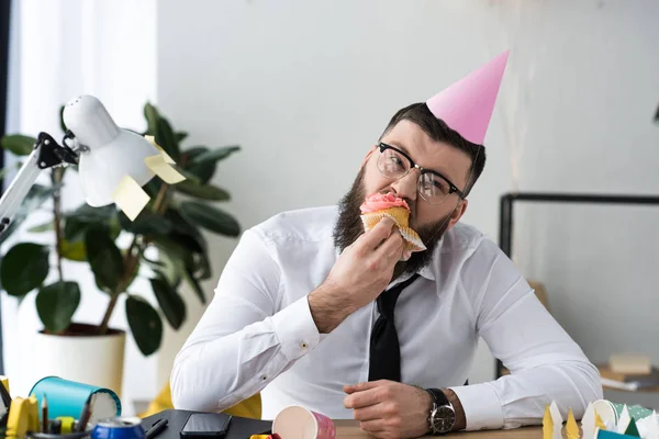 Портрет бизнесмена в конусе вечеринки, поедающего праздничный кекс в офисе — стоковое фото