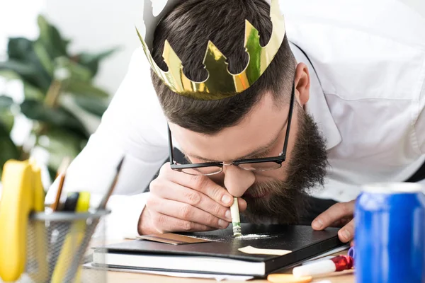 Hombre de negocios con corona de papel en la cabeza tomando drogas en el lugar de trabajo en la oficina - foto de stock