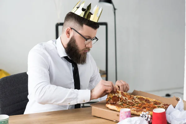 Бизнесмен в бумажной короне сидит на рабочем месте с пиццей — стоковое фото