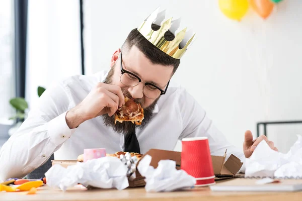 Porträt eines Geschäftsmannes mit Papierkrone auf dem Kopf, der am Arbeitsplatz Pizza isst — Stockfoto