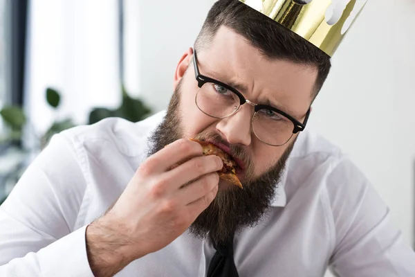 Портрет унылого бородатого бизнесмена с бумажной короной на голове, поедающего пиццу — стоковое фото