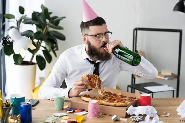 Empresário com cone de festa na cabeça bebendo champanhe enquanto come pizza no local de trabalho — Fotografia de Stock