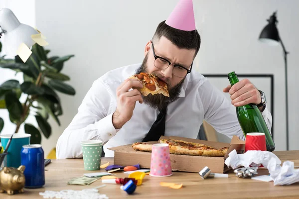 Бизнесмен с конусом вечеринки на голове ест пиццу на рабочем месте — стоковое фото