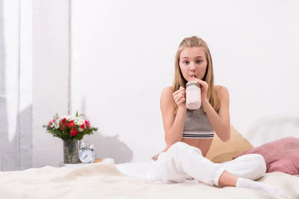 Attraktive junge Frau trinkt Milchshake während sie auf dem Bett sitzt — Stockfoto