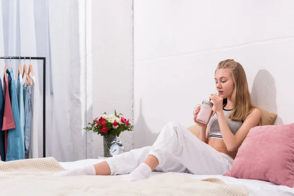 Beautiful young woman drinking milkshake while sitting on bed — Stock Photo