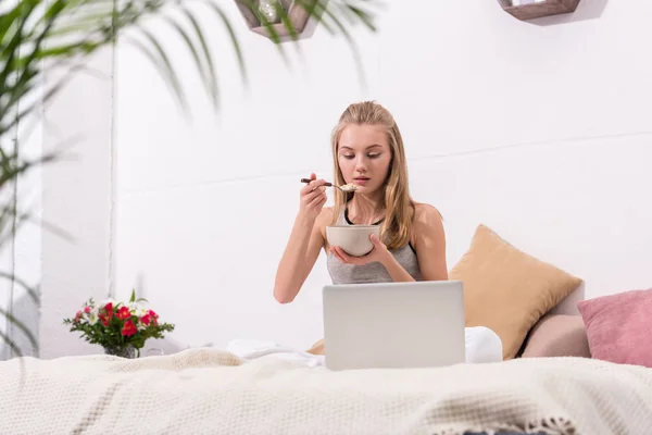 Jovem mulher comendo café da manhã de cereais na cama de manhã e olhando para laptop — Fotografia de Stock