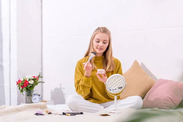 Happy young woman applying blush while sitting on bed at home — Stock Photo