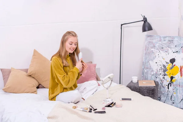 Jovem mulher escovando o cabelo enquanto sentado na cama em casa — Fotografia de Stock