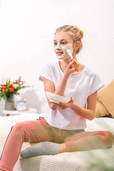Attractive woman applying clay mask on face while sitting on bed — Stock Photo