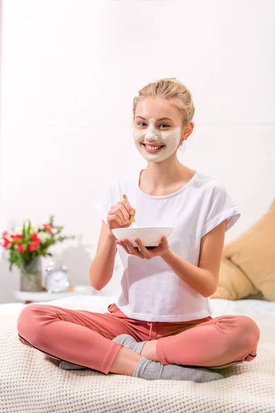 Glückliche junge Frau trägt weiße Tonmaske auf Gesicht auf, während sie auf dem Bett sitzt — Stockfoto