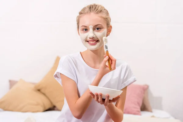 Young woman applying white clay mask on face while sitting on bed — Stock Photo