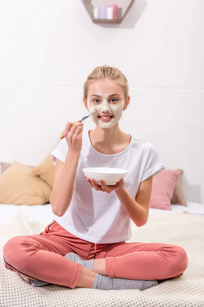 Happy young woman applying clay mask on face at home and looking at camera — Stock Photo