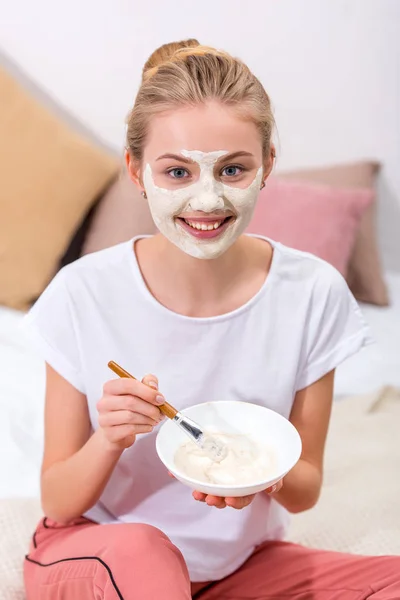 Junge Frau trägt zuhause Tonmaske auf Gesicht auf und schaut in die Kamera — Stockfoto