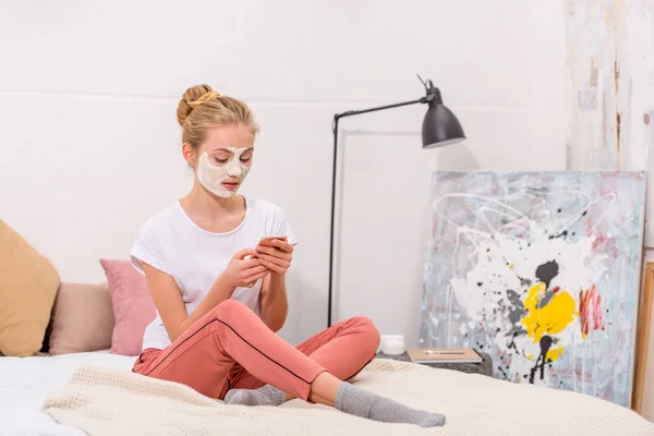 Jeune femme avec masque facial en argile blanche à l'aide d'un smartphone assis sur le lit à la maison — Photo de stock