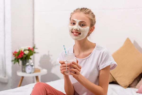 Lächelnde junge Frau mit weißer Tongesichtsmaske mit Milchshake im Einmachglas zu Hause — Stockfoto