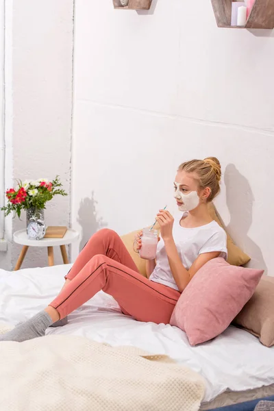 Молодая женщина в белой глиняной маске для лица пьет молочный коктейль на кровати дома — стоковое фото