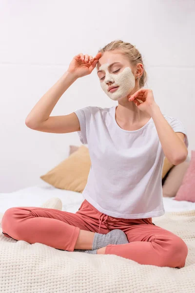 Jovem mulher com máscara facial de barro branco sentado na cama em casa — Fotografia de Stock