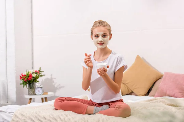 Junge Frau trägt Tonmaske auf Gesicht auf, während sie zu Hause auf dem Bett sitzt — Stockfoto