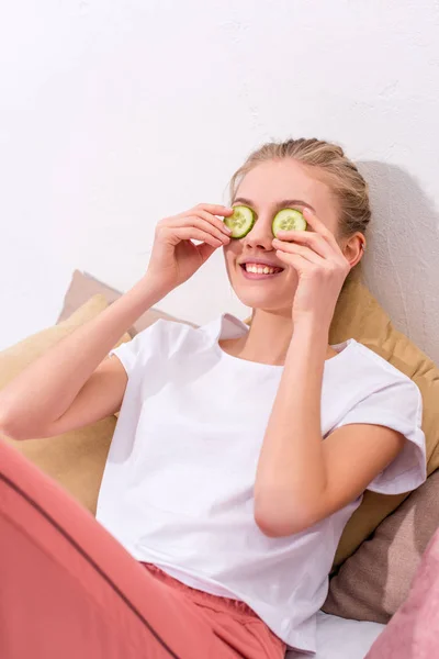 Jovem feliz aplicando fatias de pepino nos olhos em casa — Fotografia de Stock