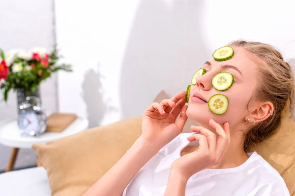 Young woman applying cucumber slices on face at home — Stock Photo