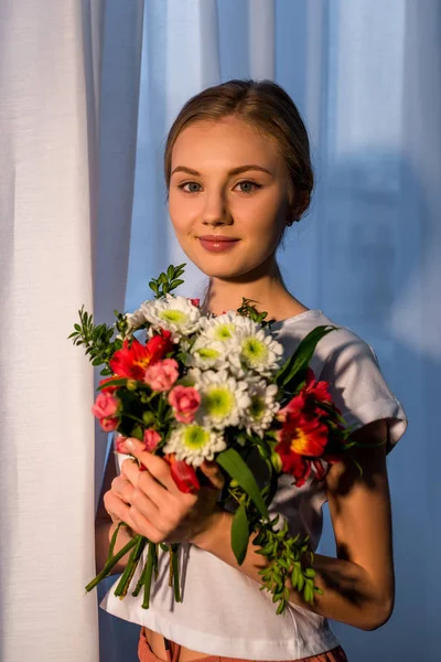 Jeune femme avec beau bouquet regardant caméra contre fenêtre — Photo de stock