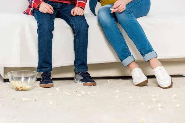 Ritagliato colpo di madre e bambino seduto sul divano con popcorn versato disordinato sul pavimento — Foto stock