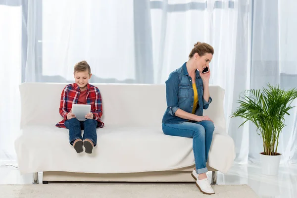 Happy little son using digital tablet while his mother talking by phone on couch — Stock Photo