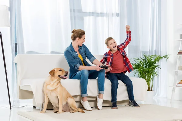 Madre y feliz pequeño hijo jugando videojuegos mientras su perro sentado en el suelo - foto de stock