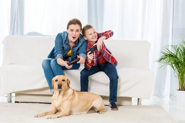 Эмоциональная мать и сын играют в видеоигры, пока их собака лежит на полу и смотрит — стоковое фото