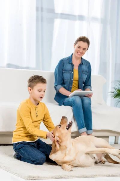 Kid petting dog on floor while mother using tablet on couch — Stock Photo