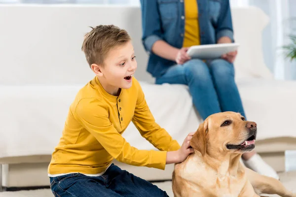 Emocionado pequeño niño acariciando perro en el suelo mientras que la madre usando tableta en el sofá - foto de stock
