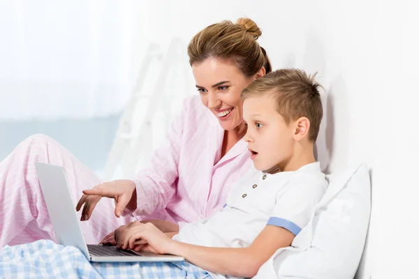 Madre feliz e hijo emocional usando el ordenador portátil juntos en la cama - foto de stock