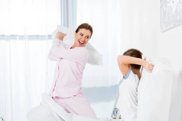 Feliz madre e hijo peleando con almohadas en la cama en la mañana del fin de semana - foto de stock