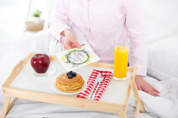 Colpo ritagliato di donna con colazione a letto e biglietto di auguri per la giornata delle madri — Foto stock