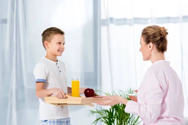 Glücklicher kleiner Sohn trägt Frühstück auf Holztablett für Mutter — Stockfoto