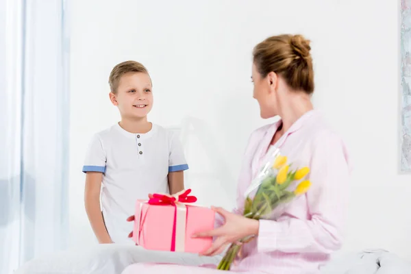 Madre con el regalo y el ramo de flores presentado por el pequeño hijo en la mañana del día de las madres - foto de stock