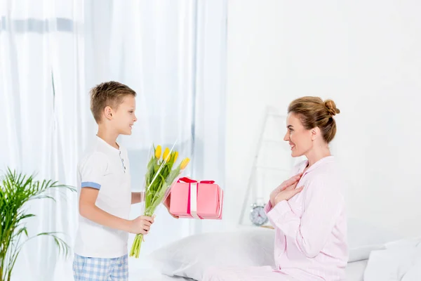 Piccolo figlio che presenta regalo e fiori per la madre il giorno delle madri mattina — Foto stock